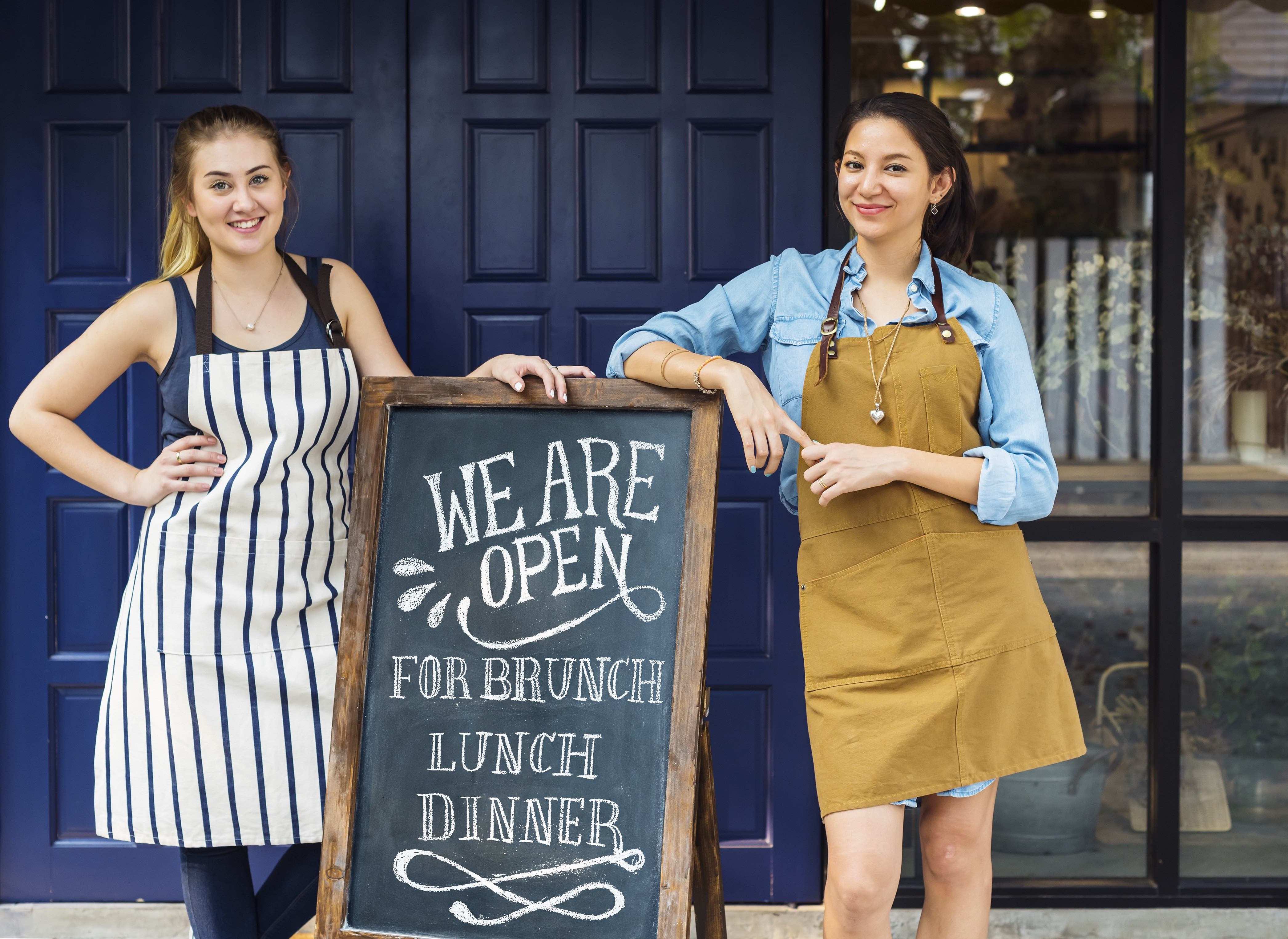 Two young woman smiling and standing in front in front of their business with a "We are open" sign in between them