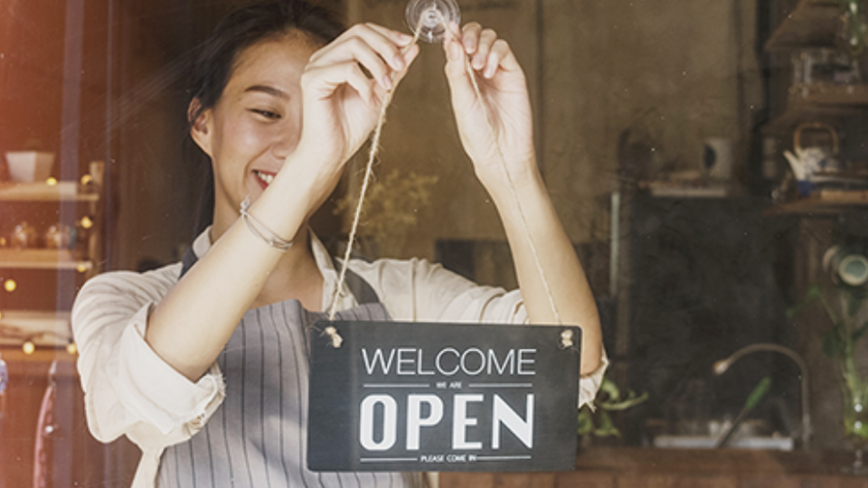 Smiling woman with dark hair holding a sign saying "Welcome we are open" at a small shop.
