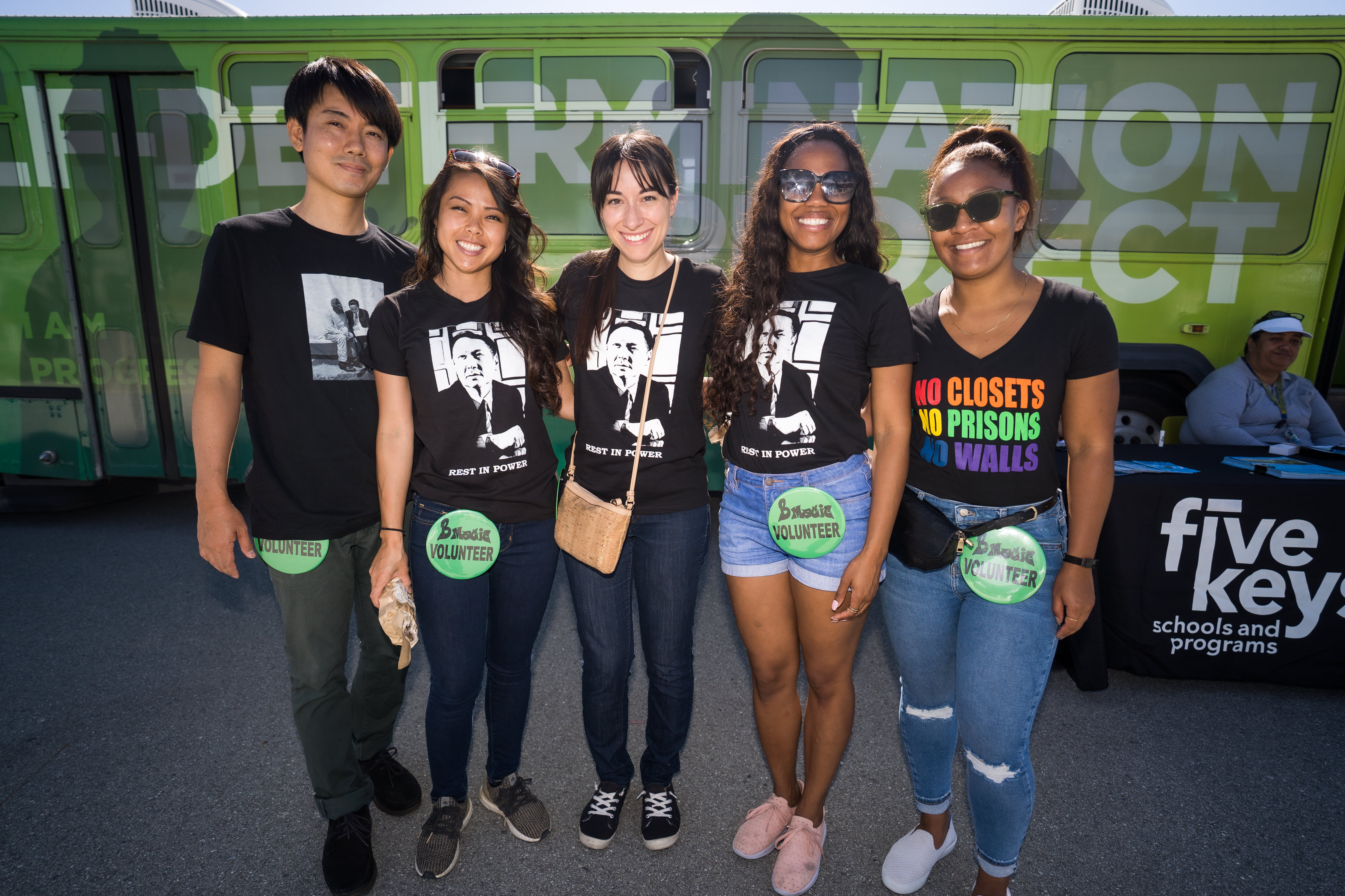 5 people stand in front of a bus. They are wearing shirts with a portrait of late Public Defender Jeff Adachi and the words "rest in power." Behind them is a table for Five Keys Schools and Programs.