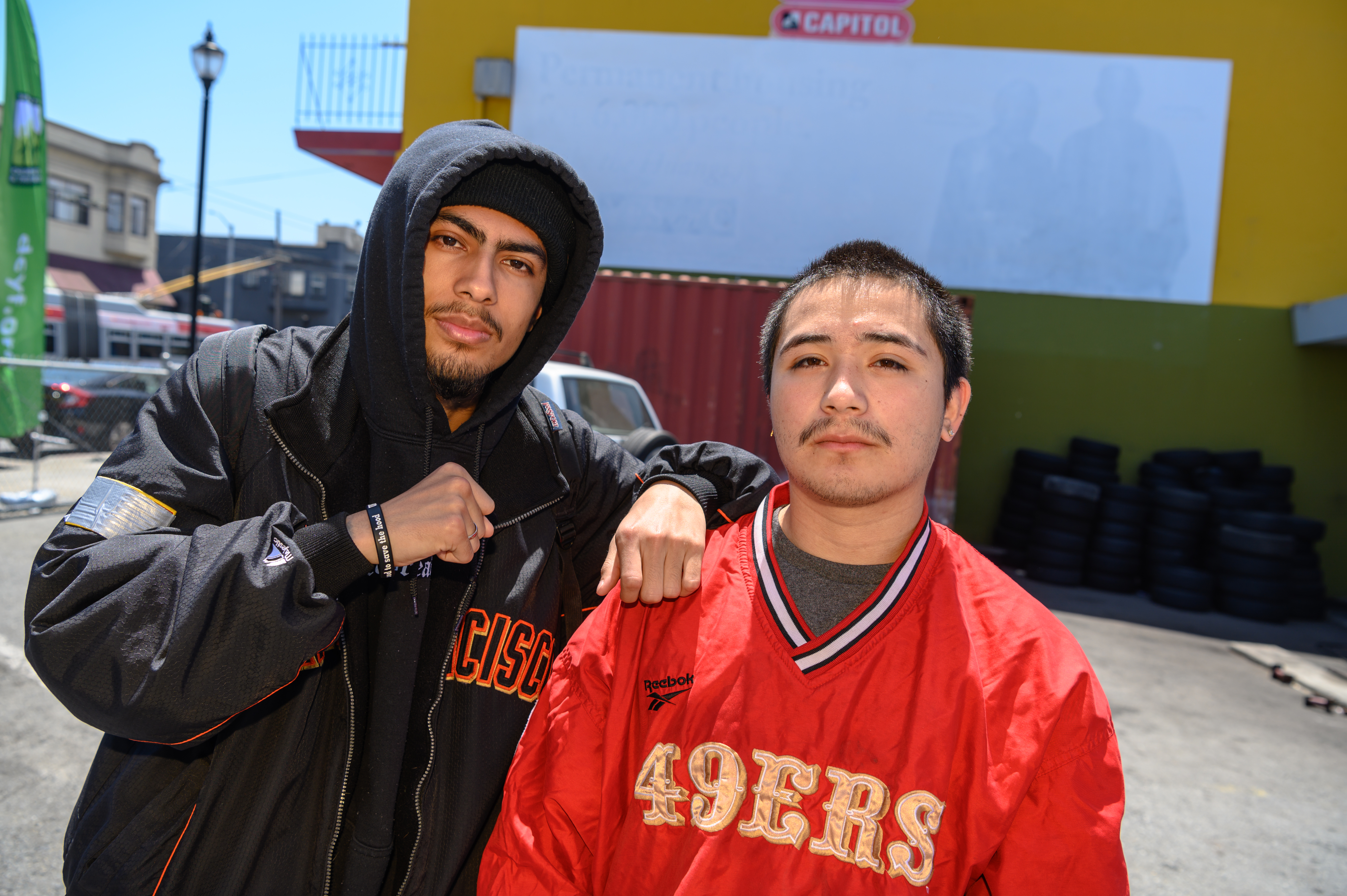 Two young men standing next to each other. The one on the left wears a black Giants jacket over a black hoodie. The one on the right wears a red 49ers pullover.