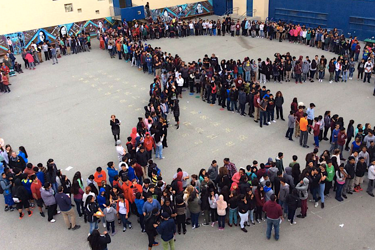 A crowd of middle schoolers in a schoolyard gathered in the shape of a peace sign
