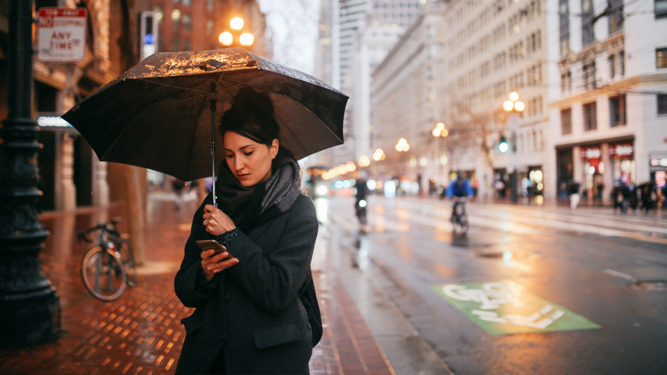 Young woman checking her smartphone while standing under an umbrella on a rainy afternoon.