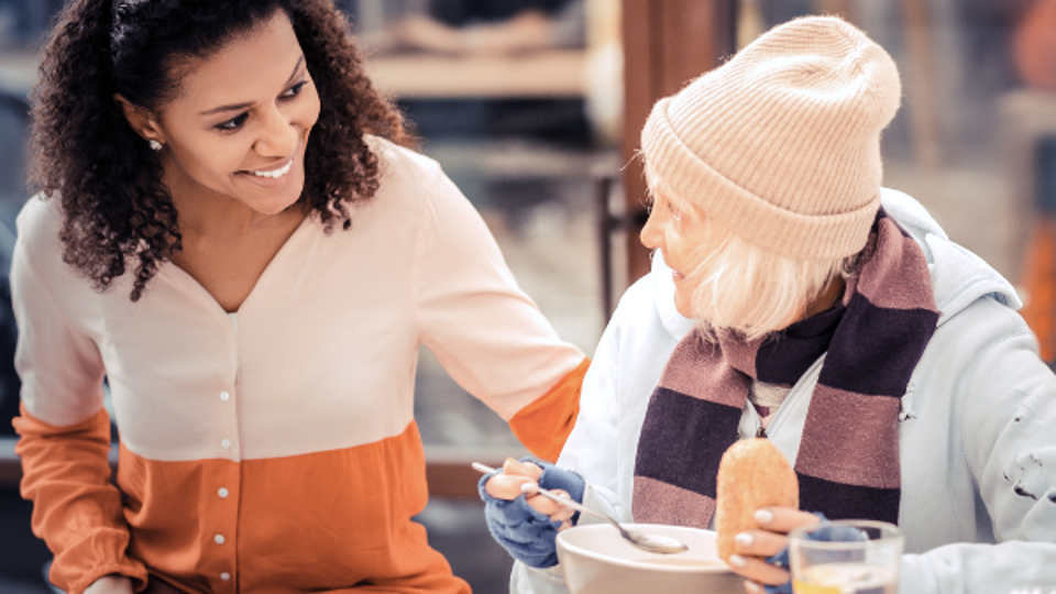 A young woman smiles at an elderly woman in need who is eating at a table.