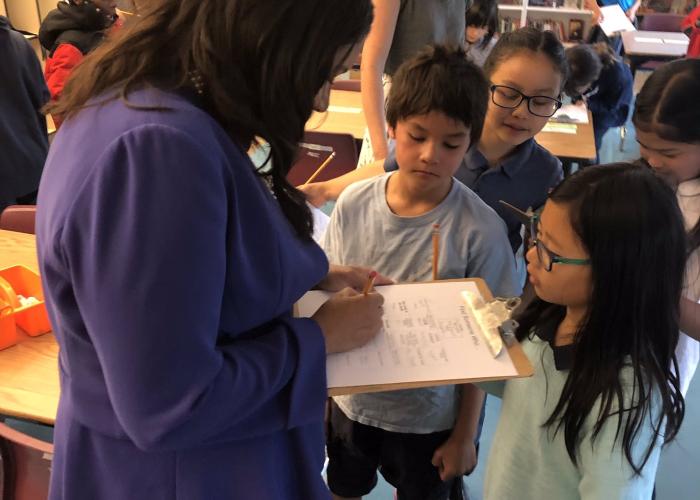 Mayor London Breed stands in a classroom using a clipboard while children surrounded by a group of curious children