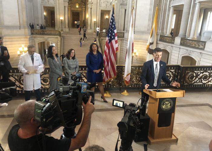 Dr. Grant Colfax speaking at a podium at a press conference with Mayor London Breed.