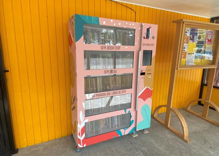 The SFPL Book Kiosk has a pink wrapper and stands in front of Island Cove Market.