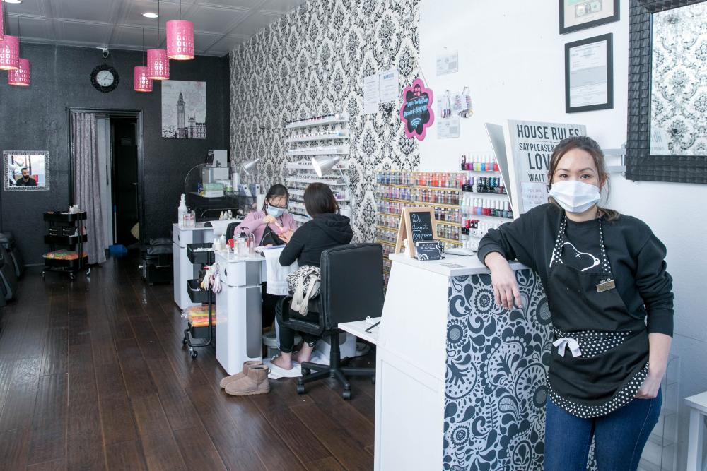Nail salon owner standing at desk wearing a mask with nail salon in the background.