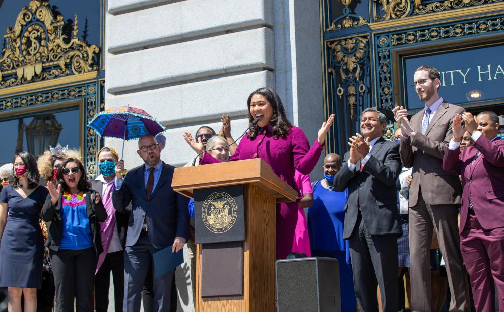 Mayor London Breed speaks at a podium on the steps of City Hall, surrounded by City officials and special guests.