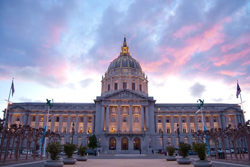 A view looking up at the lit front facade of San Francisco City Hall at dusk, with a brilliant purple and pink sky in the background.