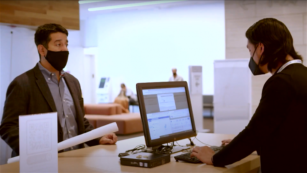 A customer holding a rolled-up plan stands at a counter while a Permit Center staff member is on the computer working on his intake. Both are masked.