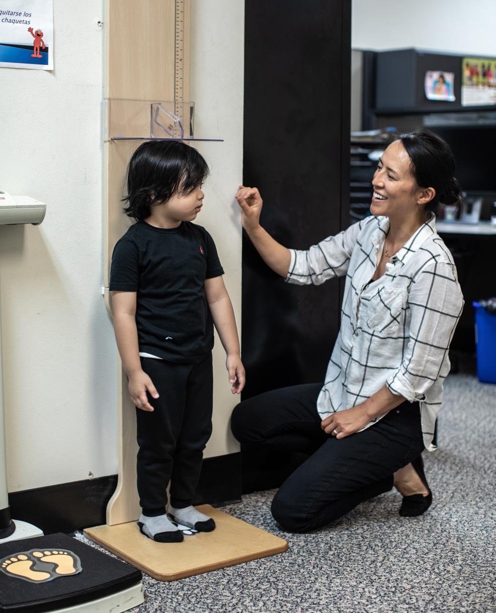 Female provider is kneeling besides a toddler boy as she measures his height using a stadiometer.