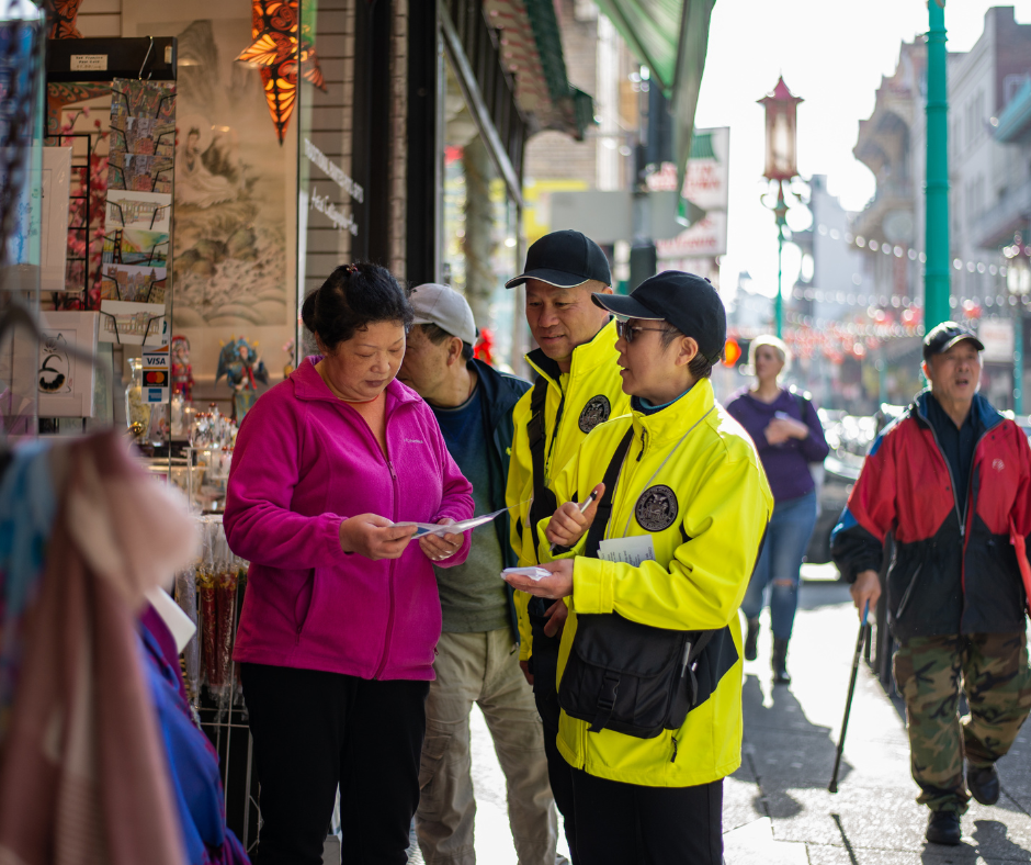 Two individuals wearing neon yellow jackets speak to a shop owner outside of their storefront in Chinatown.