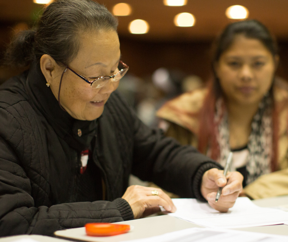 Woman smiles as she fills out her citizenship paperwork at a free workshop, while another woman looks on