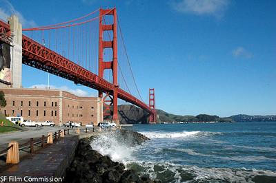 Looking up at the Golden Gate Bridge from a rocky shore, with the waves breaking on the rocks. It is a sunny day with few clouds.