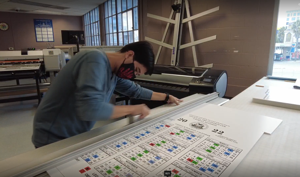 Individual uses large paper cutter to trim a payroll calendar poster.