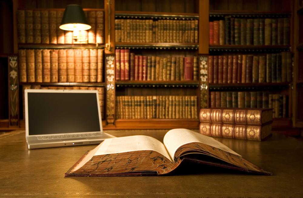 A large bound book lays open on a wooden table with an open laptop in the background, surrounded by a shelf of legal books