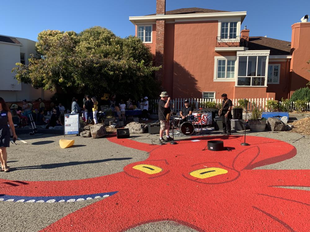 A band plays for a crowd in the middle of a public parking lot, on top of a large octopus mural.