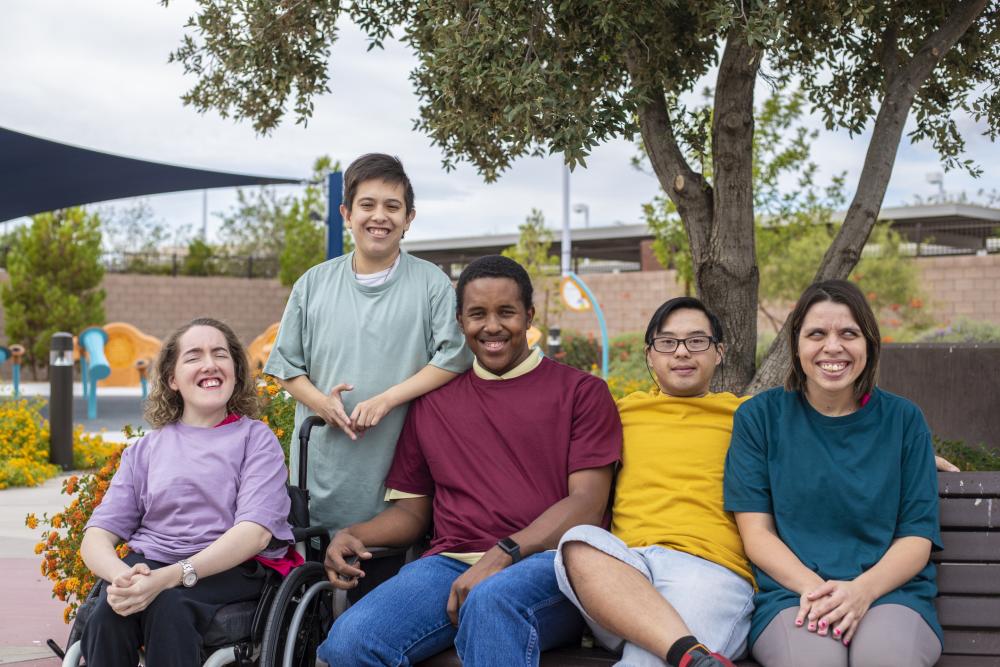 A group of people with disabilities. One person is standing, three people are sitting on a bench, and one person is sitting on a wheelchair. 