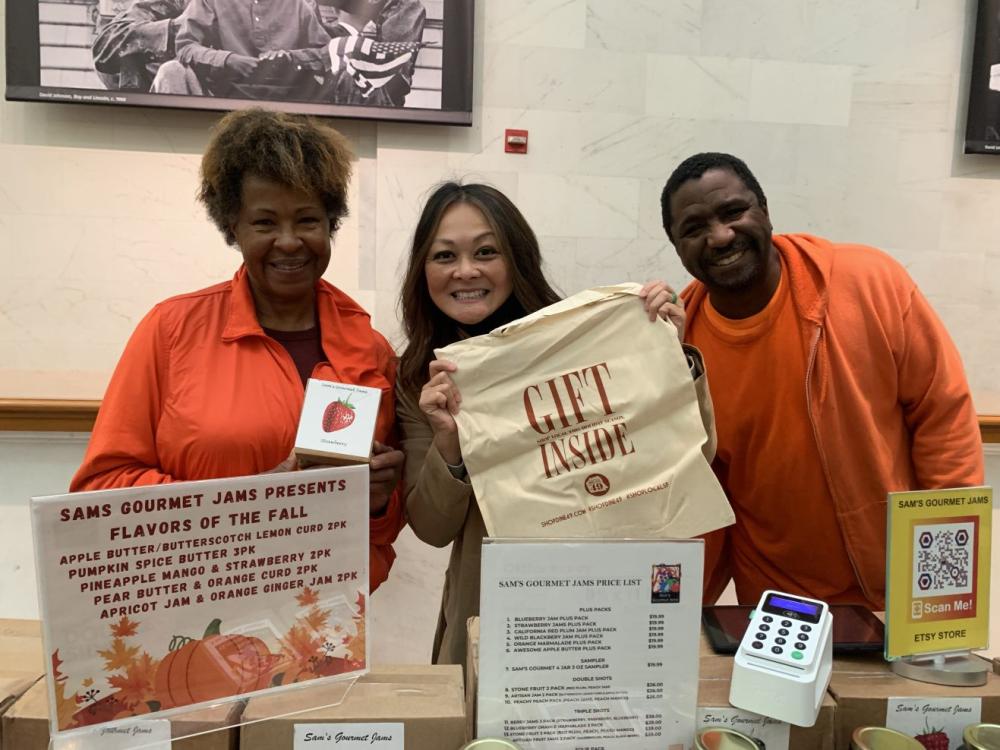 City Administrator Carmen Chu and vendors pose behind a display selling jams. City Administrator Chu holds up a tote bag that says "Gift Inside!"