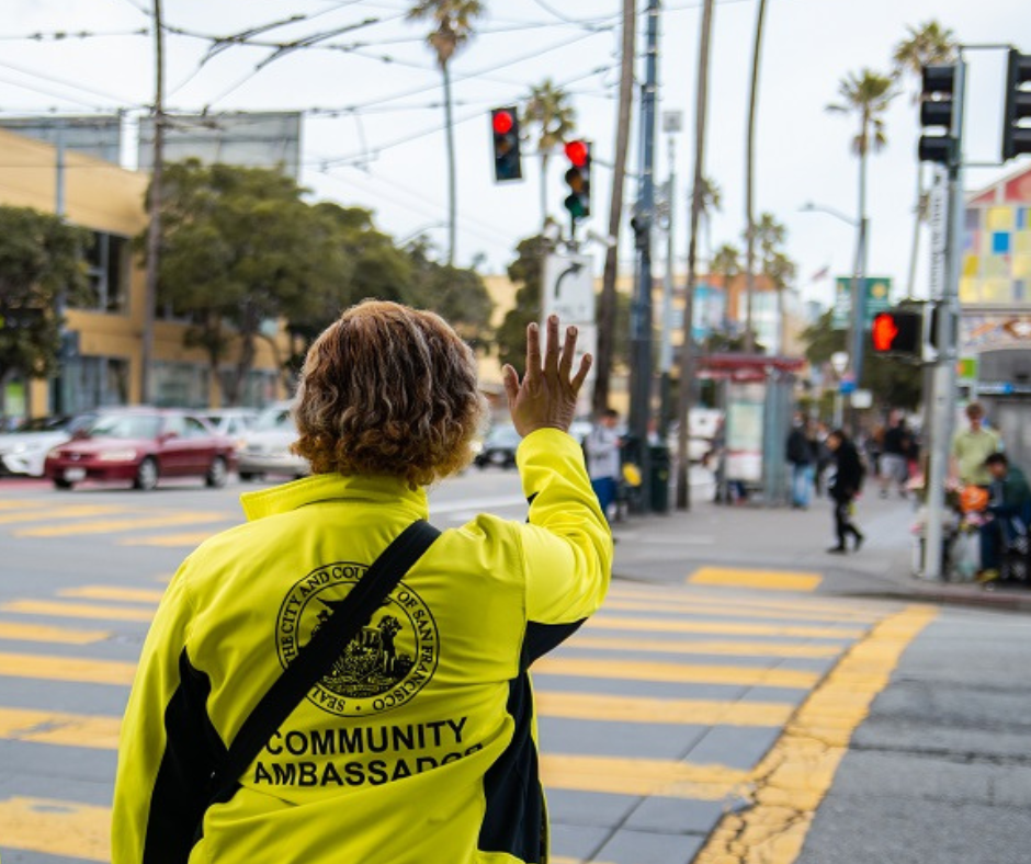 A Community Ambassador in a yellow jacket stands at the intersection of 16th and Mission streets and waves