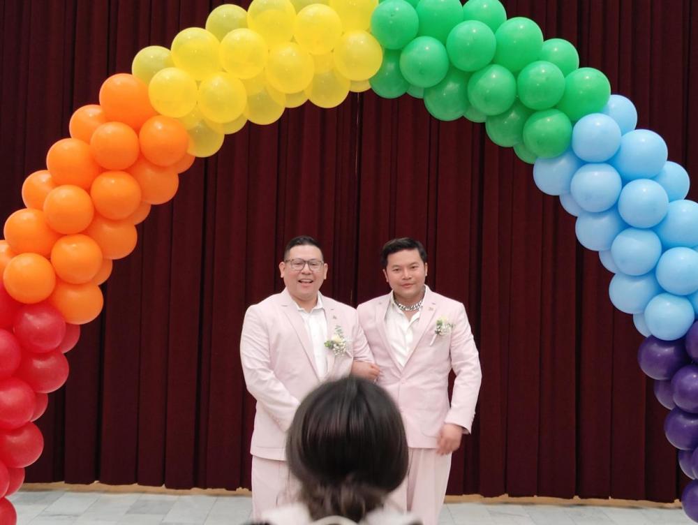 Two individuals in matching light pink suits stand together under a vibrant, rainbow-colored balloon arch. They both wear boutonnieres, and one has his arm gently resting on the other’s. The background is a rich red curtain, and a person in the foreground partially appears, facing away from the camera. The scene has a celebratory and joyful atmosphere.