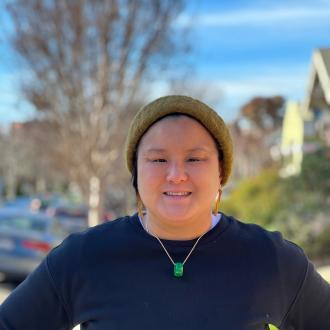 impage of person wearing a brown beanie and dark sweater with a necklace and earrings, smiling in a sunny green background