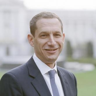 Daniel Lurie, 46th Mayor of San Francisco, in a dark suit and tie is standing outdoors in front of a green lawn and a blurred City Hall in the background.